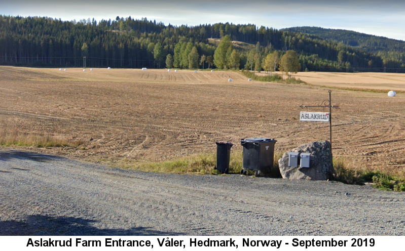 Aslakrud Farm Entrance Våler, Hedmark, Norway: Color photo, taken in September 2019, of a grey gravel road leading away from a wooden sign, hanging from a wrought-iron post, that reads 'Aslakrud'; behind them are brown barren fields lying before a wooded hillside